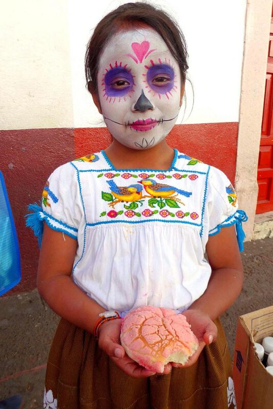 Little girl in Catrina makeup holds a loaf of “pan de muerto,” the traditional sweet bread baked for the Day of the Dead.